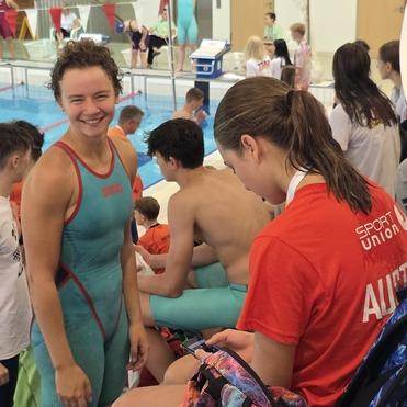 Junge Schwimmerin in Badeanzug lächelt am Beckenrand, während andere in Badebekleidung sitzen und sich vorbereiten.