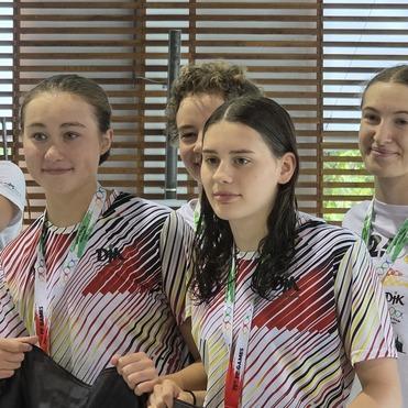 Gruppe junger Schwimmerinnen mit nassen Haaren, die in Sportbekleidung an einem Holzpaneel stehen.
