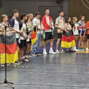 Gruppierung von jugendlichen Sportlern in verschiedenen Trikots mit Medaillen auf einer Sportveranstaltung, während sie applaudiert werden.