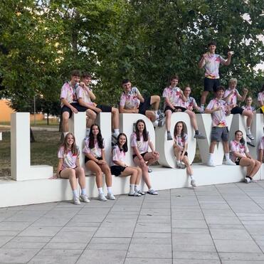 Gruppe junger Menschen sitzt und steht um ein großes, dekoratives #LLN030-Schild auf einem Stadtplatz.