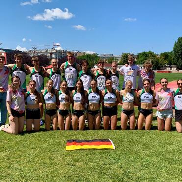 Gruppe junger Athleten in Sportkleidung posiert lächelnd auf einer Wiese vor einem Sportgelände mit Flagge Deutschlands.