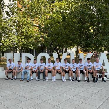 Gruppenspieler in weißen T-Shirts sitzen auf einem großen Schriftzug „L'UDOVIKA CAMPUS“ vor grüner Bepflanzung.