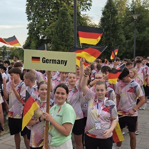 Gruppe junger Menschen mit Deutschlandfahnen, eine Person h&auml;lt ein Schild mit &bdquo;Germany&ldquo; und alle tragen einheitliche T-Shirts.