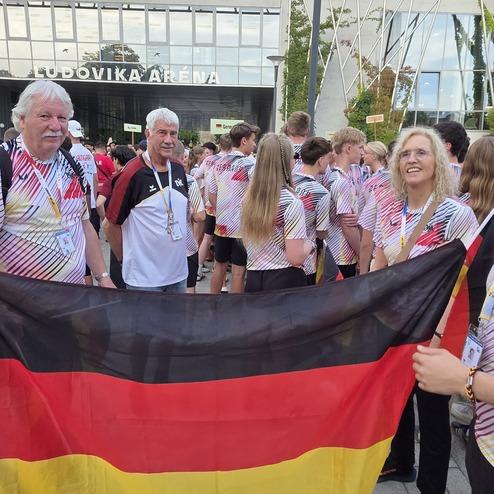 Gruppe von f&uuml;nf Personen in bunten Trikots vor einem Geb&auml;ude, halten eine deutsche Flagge und stehen in einer Menschenmenge.