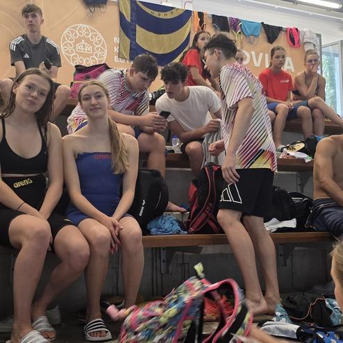 Jugendliche schwimmen und sitzen in einem Schwimmhalle auf Trib&uuml;nen, einige tragen Badebekleidung, andere sportliche Outfits.