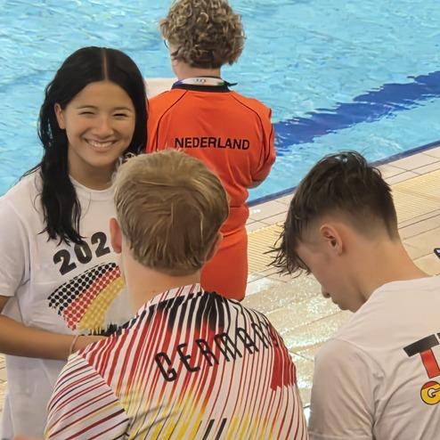 L&auml;chelnde Sportlerin in Mannschaftsanzug spricht mit zwei anderen Athleten am Schwimmbecken. Hintergrund zeigt Zuschauer und weitere Teilnehmer.