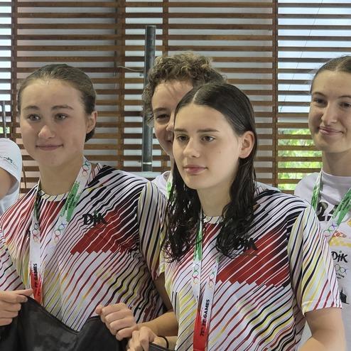 Gruppe junger Schwimmerinnen mit nassen Haaren, die in Sportbekleidung an einem Holzpaneel stehen.