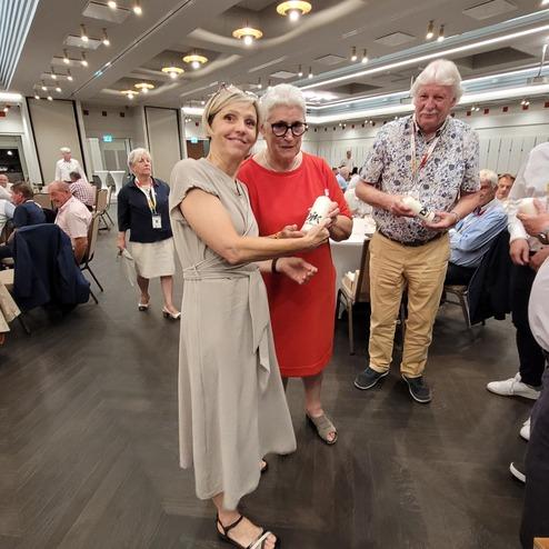 Zwei Frauen tauschen Geschenke aus, umgeben von G&auml;sten bei einer Feier in einem modernen Veranstaltungsraum.