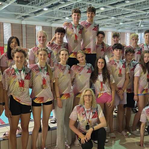 Gruppe von Schwimmern in bunten Trikots steht am Beckenrand eines Schwimmbads und l&auml;chelt in die Kamera.