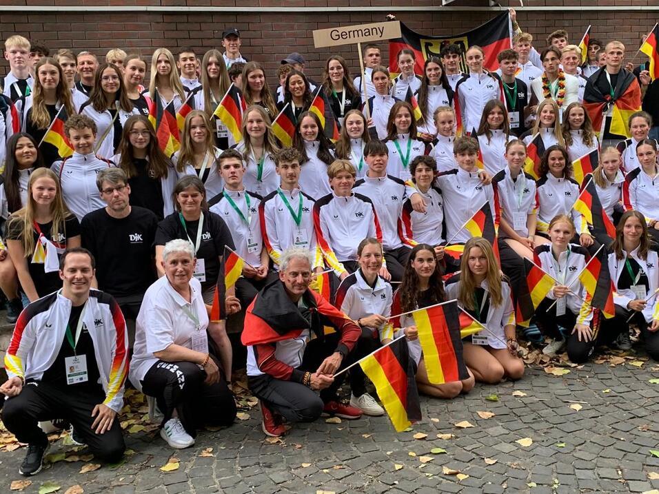 Gruppenfoto von Sportlern in weißen und schwarzen Trikots, die deutsche Flaggen vor einem Gebäude halten.