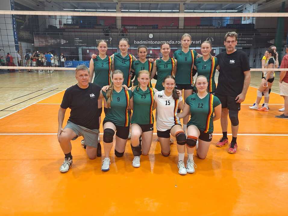 Gruppenteilnehmer in Sporttrikots stehen vor einem Volleyballnetz in einer Halle, zwei Trainer knien links und rechts.