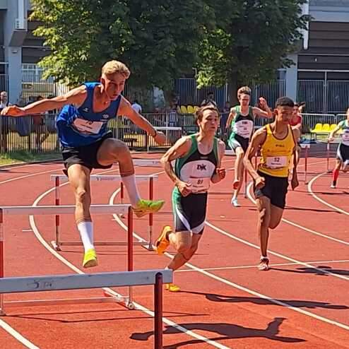 Zwei Athleten sprinten auf einer Leichtathletikbahn, einer &uuml;berspringt eine H&uuml;rde, w&auml;hrend der andere daneben l&auml;uft.