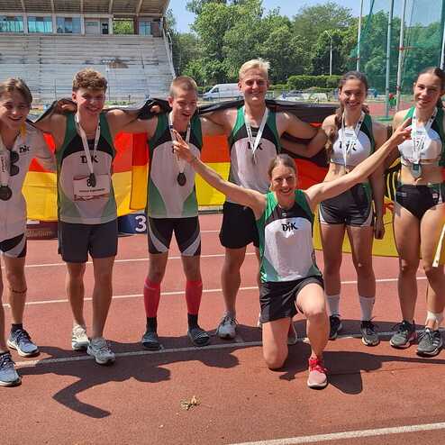 Gruppenselfie von sieben Leichtathleten mit Medaillen, umgeben von Deutschlandflaggen auf einem Sportplatz.