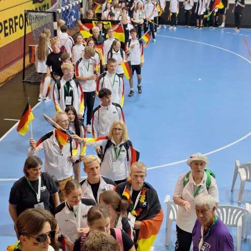 Gruppierung von Sportler*innen in Team-Outfits mit deutschen Fahnen beim Einmarsch in eine Sporthalle.