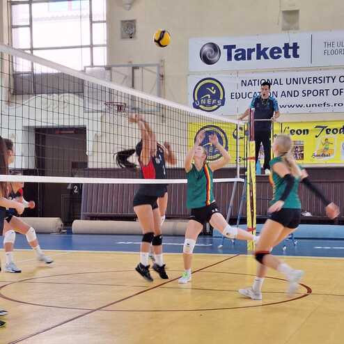 Spielszene in der Volleyball-Halle mit mehreren Spielerinnen in Aktion, w&auml;hrend ein Ball &uuml;ber das Netz fliegt.