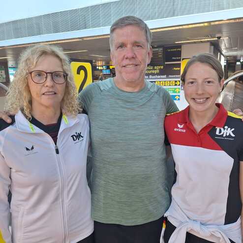 Drei Personen stehen l&auml;chelnd in einem modernen Flughafen, im Hintergrund sind Anzeigetafeln sichtbar.