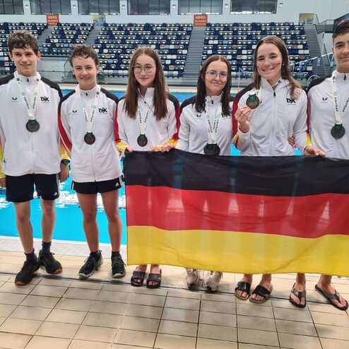 Sechs Schwimmer in wei&szlig;en Jacken mit Medaillen halten eine deutsche Flagge vor einer Schwimmhalle mit Beckeneinblick.