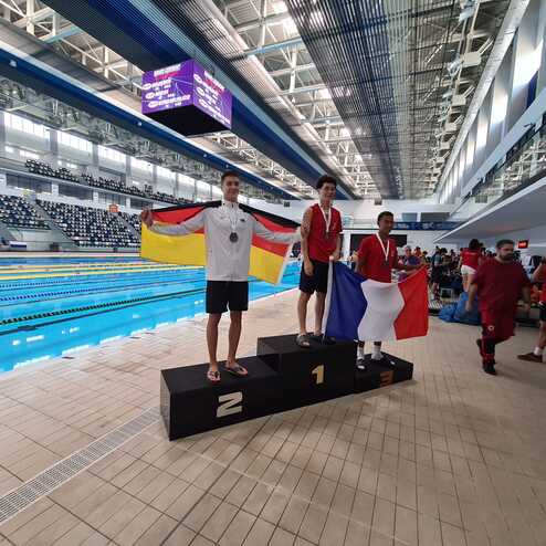 Drei Medaillengewinner stehen auf einem Siegerpodest am Schwimmbecken, einer mit deutscher, zwei mit franz&ouml;sischer Flagge.