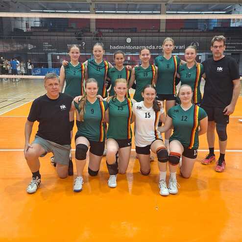 Gruppenteilnehmer in Sporttrikots stehen vor einem Volleyballnetz in einer Halle, zwei Trainer knien links und rechts.