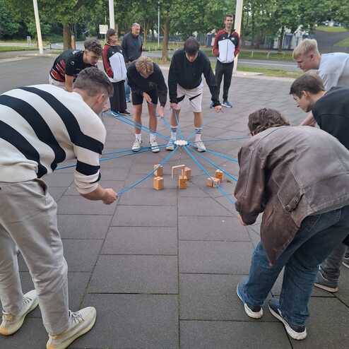 Gruppe von Jugendlichen, die sich um aufgestellte Beh&auml;lter beugen, w&auml;hrend sie Aufgaben auf dem Gehweg l&ouml;sen.