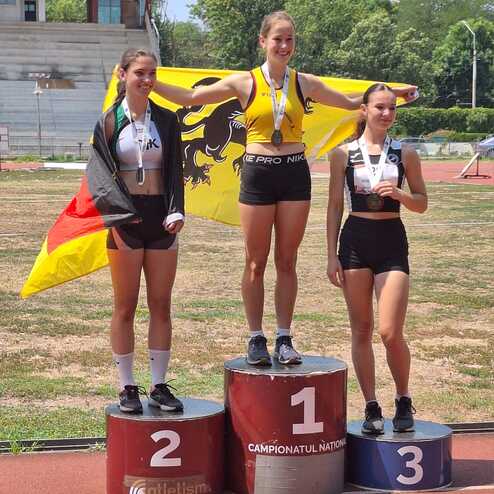 Drei Athletinnen stehen auf dem Siegerpodest; die Gewinnerin h&auml;lt eine Fahne und tr&auml;gt eine Medaille.