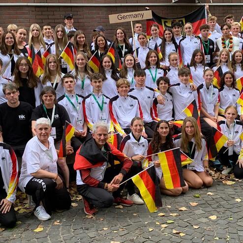 Gruppenfoto von Sportlern in wei&szlig;en und schwarzen Trikots, die deutsche Flaggen vor einem Geb&auml;ude halten.