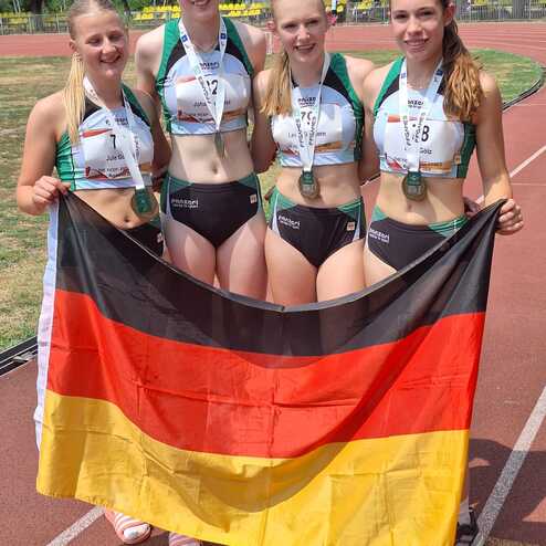 Vier junge Athletinnen in Sportkleidung halten eine deutsche Flagge und zeigen ihre Medaillen auf einer Laufbahn.