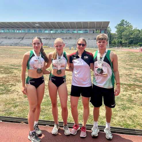 Vier Athleten mit Medaillen stehen auf einem Sportplatz, hinter ihnen ist ein leeres Stadion mit Trib&uuml;nen sichtbar.