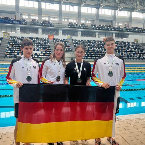 Vier Schwimmer in Sportbekleidung halten eine deutsche Flagge und zeigen ihre Medaillen neben einem Schwimmbecken.