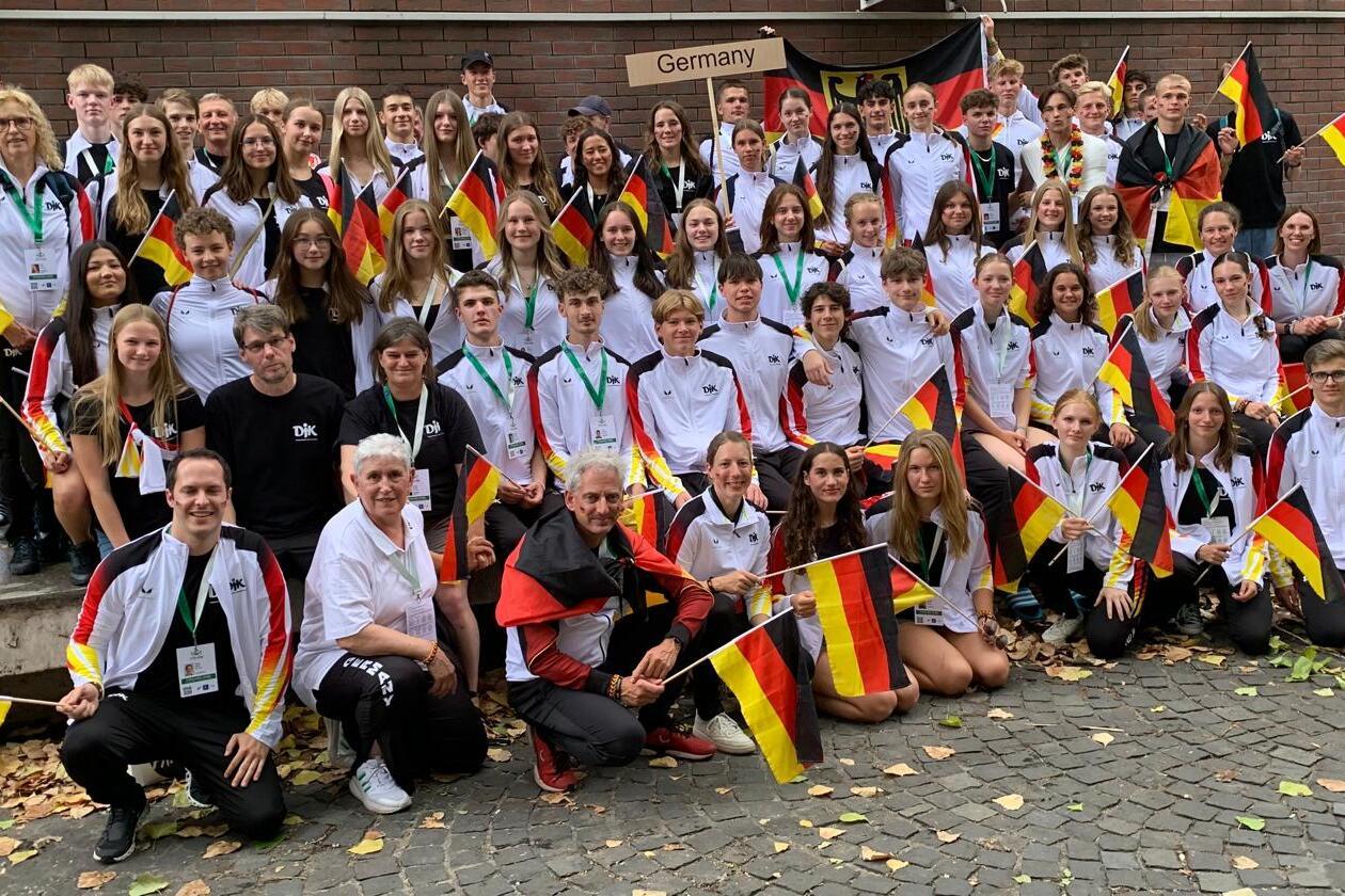 Gruppenfoto von Sportlern in wei&szlig;en und schwarzen Trikots, die deutsche Flaggen vor einem Geb&auml;ude halten.