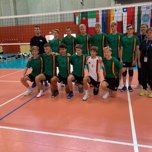 Gruppennaufnahme von 14 Volleyballspielern in gr&uuml;nen Shirts und einem Trainer auf einem bunten Volleyballfeld.