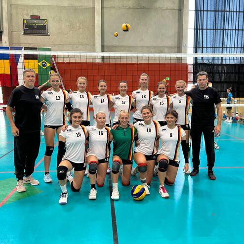 Gruppenfoto eines Volleyballteams mit 14 Spielerinnen und Trainern in einer Sporthalle, im Hintergrund ein Volleyballnetz.
