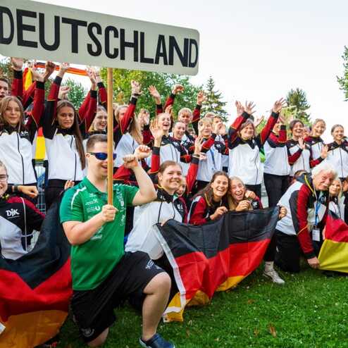 Gruppenzusammenkunft von Sportlern in Trainingsjacken mit Deutschland-Schild und Fahnen im Freien.