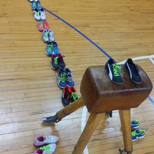 Anordnung von Sportschuhen in einer Linie neben einem Turnpferd auf einem Holzfu&szlig;boden in einer Sporthalle.