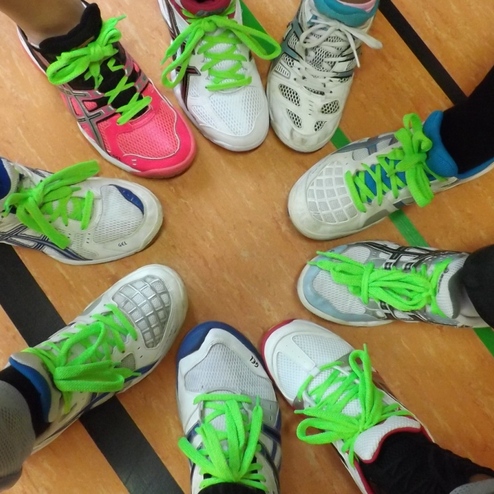 Ansicht von acht F&uuml;&szlig;en in Sportschuhen mit leuchtend gr&uuml;nen Schn&uuml;rsenkeln, umringt von einem Holzfu&szlig;boden.