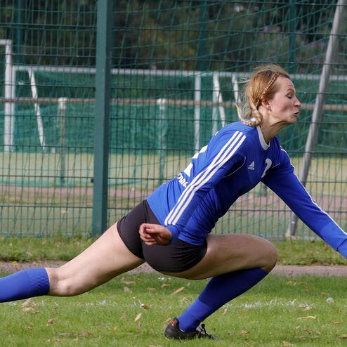 Sportlerin in blauer Trikot und Shorts weicht einem Ball aus, während sie sich auf einem Fußballfeld in der Luft streckt.