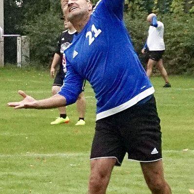 Ein Mann wirft einen Volleyball über ein Netz auf einem Sportplatz, während andere Spieler im Hintergrund stehen.