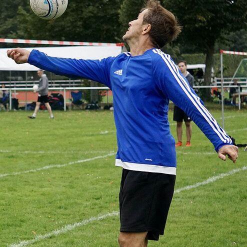 Mann in blauer Sportjacke hebt einen Ball auf einem Fußballfeld mit grüner Wiese zum Wurf an.