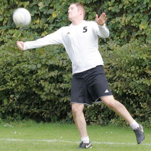 Ein Spieler in weißem Trikot wirft einen Ball auf einem Fußballfeld mit grünem Hintergrund.