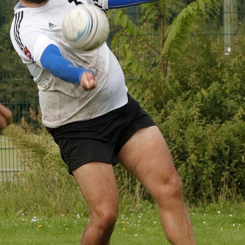 Mann in Sportkleidung kickt einen Ball auf einer grünen Wiese, umgeben von Büschen und einem Zaun im Hintergrund.