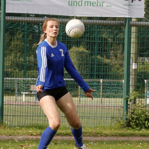 Eine junge Frau in blauer Sportkleidung spielt Fußball und hebt den Ball über ihre Schulter auf dem Platz.