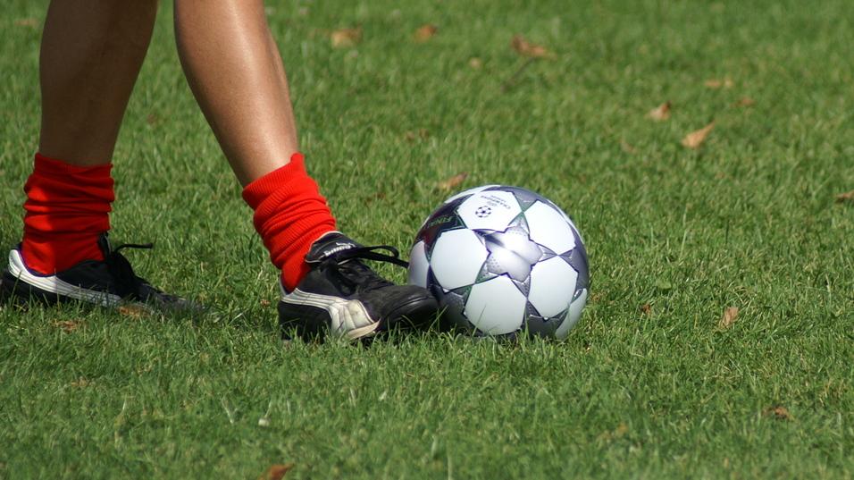 Nahaufnahme eines Fußes in schwarzen Schuhen mit roten Socken, der einen Fußball auf grüner Wiese berührt.