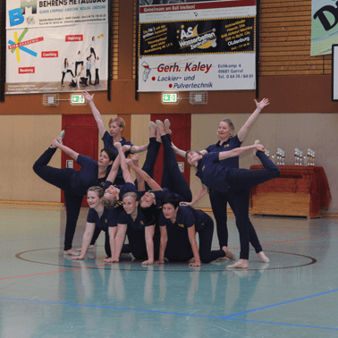 Gruppe von neun T&auml;nzerinnen in Sportkleidung zeigt eine Pose auf einem Hallenboden mit bunten Sponsorenplakaten.