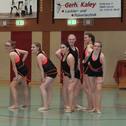 Sechs T&auml;nzerinnen in Schwarz und Pink stehen in einer choreografierten Pose auf einem Sportboden.