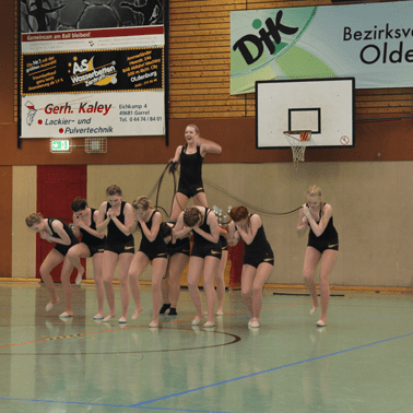 Eine Gruppe von M&auml;dchen in schwarzen Trikots f&uuml;hrt eine synchronisierte Gymnastik&uuml;bung in einer Sporthalle aus.