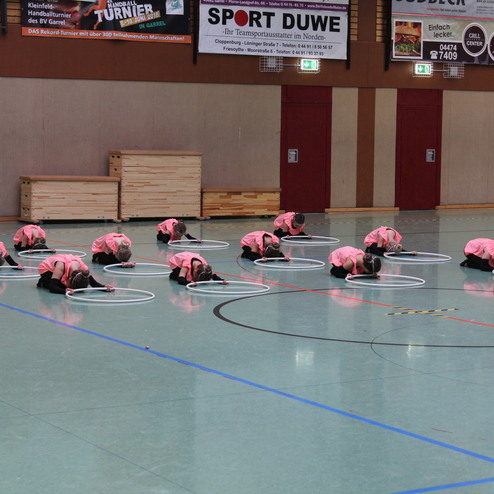 Gruppen von M&auml;dchen in pinken Shirts &uuml;ben mit Hula-Hoops in einer Sporthalle auf gr&uuml;nem Boden.