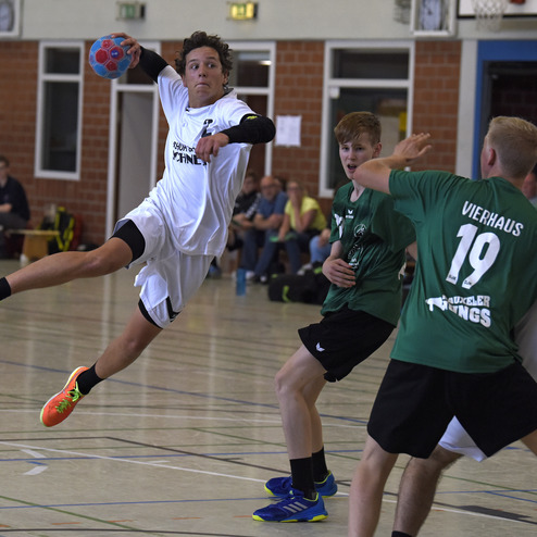 Handballspieler in Wei&szlig; springt zum Wurf, w&auml;hrend er von zwei Verteidigern in Gr&uuml;ner Trikots umgeben ist.