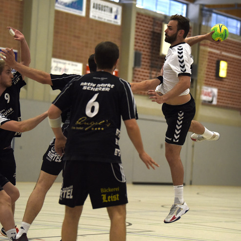 Handballspieler springt hoch, um einen Ball zu werfen, w&auml;hrend Mitspieler und Gegner ihn umringen.