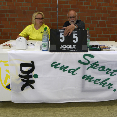 Zwei Personen sitzen an einem Tisch mit einem Scoreboard f&uuml;r ein Tischtennisturnier und einem Banner mit der Aufschrift &bdquo;Sport und mehr...&ldquo;.