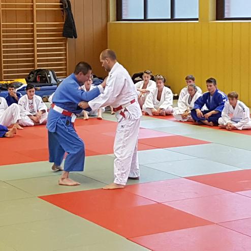 Zwei Judoka sparren auf einer Mattenfl&auml;che, w&auml;hrend eine Gruppe Zuschauer in Judogis zusieht.